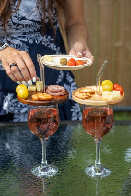 Deux couvercles d’apéro en érable et en frêne torréfié posés sur des coupes à vin rosé, garnis de fromages, charcuteries et craquelins, avec pailles en verre.