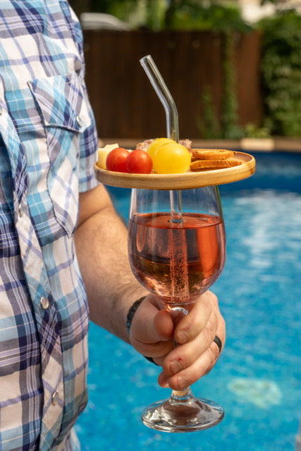 Couvercle d’apéro en érable garni de bouchées sur une coupe de vin rosé tenue d’une main, avec une paille en verre, au bord d’une piscine.