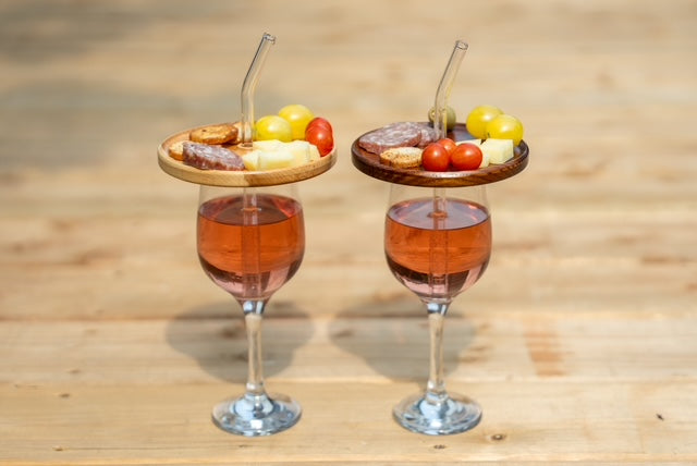 Couvercles d’apéro en érable et en frêne torréfié garnis de charcuteries, fromages et tomates, sur deux coupes de vin rosé avec pailles en verre.