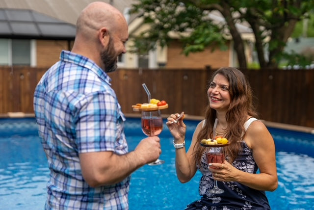 Deux personnes au bord d’une piscine utilisent des couvercles d’apéro en érable garnis de bouchées sur leurs coupes à vin, gardant une main libre.